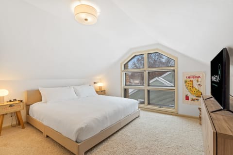 A bright attic bedroom with a large bed, sloped ceiling, and angular window, decorated with a California poster.