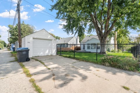 A view of a suburban alley with a white garage, trash bin, and houses behind a chain-link fence under a blue sky.