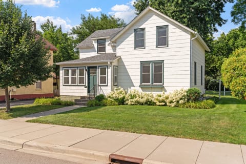 Two-story house with white siding, green accents, and flowering bushes in front.