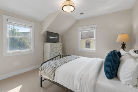 Bright bedroom featuring a bed with white linens and a blue decorative pillow, a dresser with a television, and two windows.