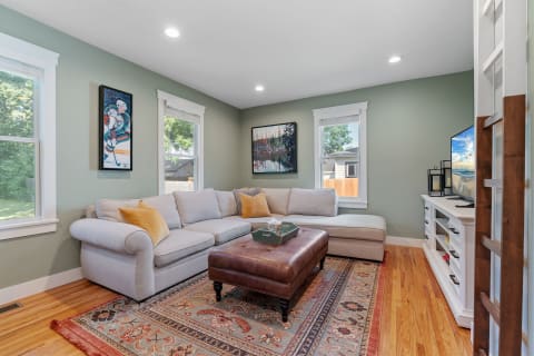 A bright living room featuring a gray sectional couch, a leather ottoman, artwork on the walls, and a television on the media console.