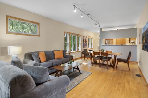 Living room with gray sofa, orange pillows, wooden dining table, and bright windows.