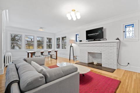 Bright living room featuring a gray sectional sofa, wooden dining table, and white-brick fireplace.