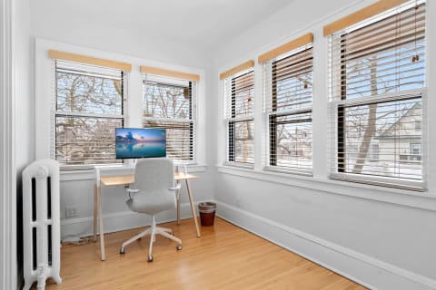 A well-lit office space featuring a wooden desk with a monitor, a gray chair, and large windows with white blinds.