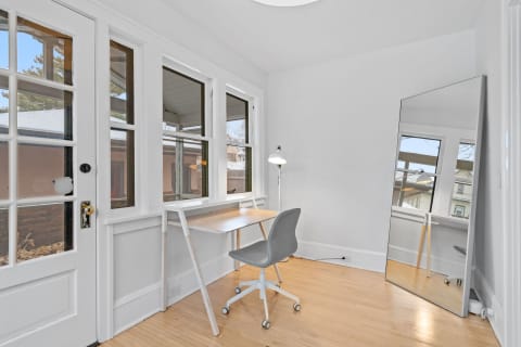 A minimalist office space featuring a wooden desk, gray chair, and large mirror, illuminated by natural light.