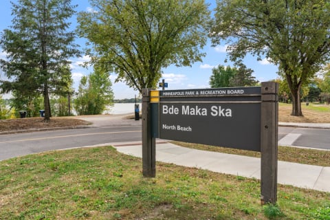 Sign marking Bde Maka Ska North Beach near a lake with trees and pathways.