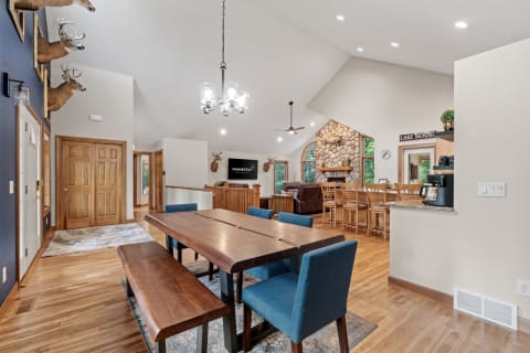 Spacious living room with wooden dining table, blue chairs, stone fireplace, and vaulted ceiling.