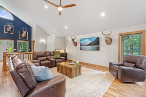 A spacious living room with leather furniture, mounted deer heads, and a television displaying a scenic landscape.