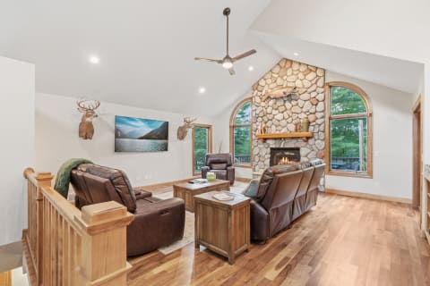 A living room featuring a stone fireplace, leather chairs, and a large TV with a view of nature outside.