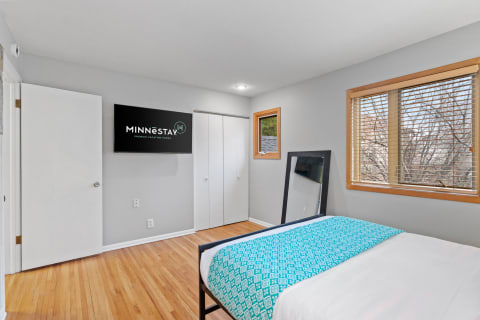 A cozy bedroom with a bed, turquoise blanket, large mirror, and a television displaying the MINNESTAY logo.