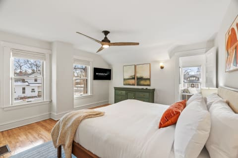 A cozy bedroom with large windows, white linens, and a green dresser, showcasing a vibrant decorative pillow.