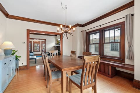 A well-designed dining room featuring a wooden table, chairs, modern chandelier, and large windows with gray curtains.
