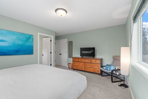 Interior view of a cozy bedroom with light green walls, a white bed, a wooden dresser, and a TV showing 'MINNESTAY'.