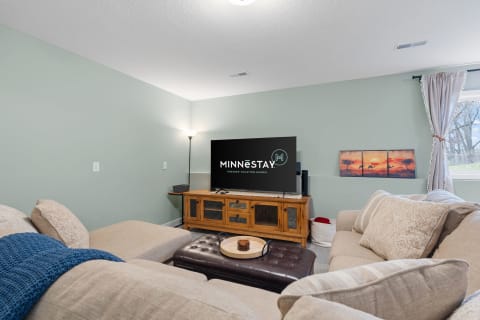 A cozy living room with a sectional couch, leather ottoman, and a wall-mounted television displaying the MINNéSTAY logo.