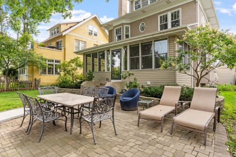 A backyard patio featuring a dining table with chairs, lounger chairs, and blooming greenery.