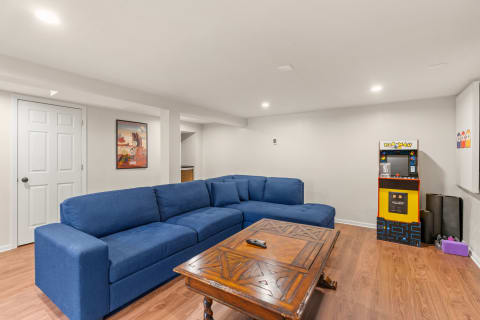 Basement living area with a blue sectional sofa, wooden coffee table, and a Pac-Man arcade machine.