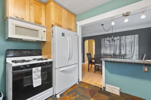 A kitchen featuring a black stove, white refrigerator, wood cabinets, and a cozy dining area with a forest-themed artwork.