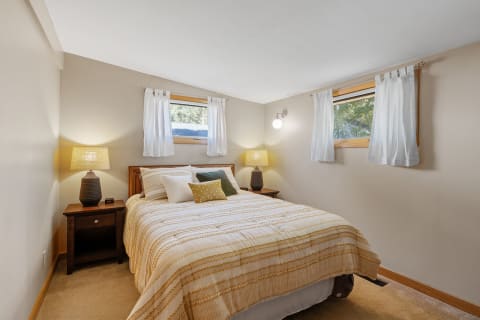 A cozy bedroom featuring a bed with decorative pillows, a bedside table with a lamp, and natural light coming from windows.