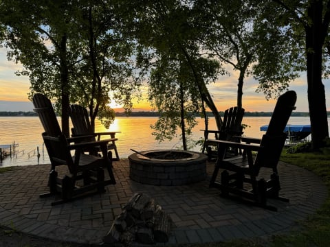 Silhouettes of rocking chairs around a fire pit by a lake at sunset.