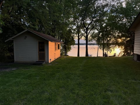 Yellow cabin by a lake at sunset with chairs on the shore.