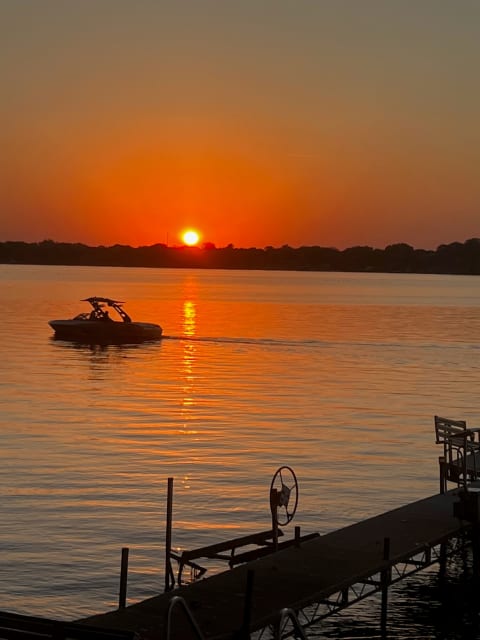 A sunset with an orange sky reflecting on a calm lake, and a boat on the water near a dock.