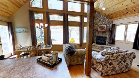 Interior of a cabin featuring a spacious living area with floral couches and a dining table, large windows overlooking a lake.