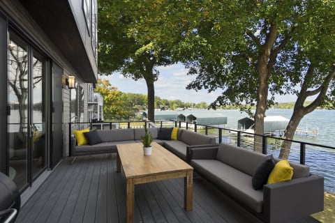 An outdoor deck with gray sofas, yellow pillows, a wooden table, and a view of a lake and trees.