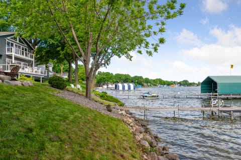 Lakeside view with a gray house, green trees, and colorful boat covers.