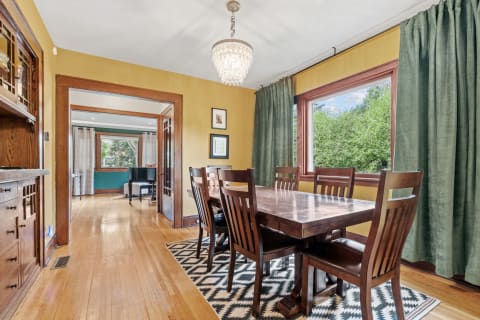 A cozy dining room featuring a wooden table, chairs, and a chandelier, with large windows and a view of greenery.