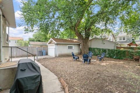 A backyard with a large tree, a seating arrangement of blue chairs, and a grill on a path.