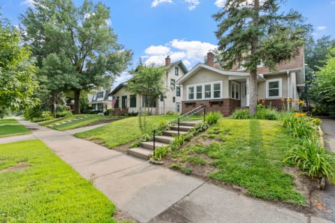 Two houses in a residential area with green lawns and blooming flowers under a blue sky.