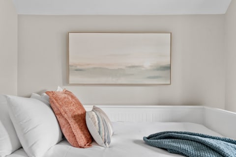 Cozy bedroom with white pillows, orange and patterned cushions, a blue blanket, and an abstract painting in soft colors.
