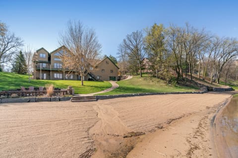 Lakeside house with sandy beach and green surroundings.