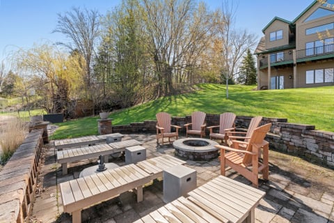 Outdoor gathering area with wooden chairs and fire pit surrounded by trees and a house.