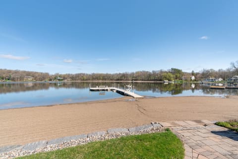 Tranquil lakeside scene with a sandy beach and wooden dock under a blue sky.