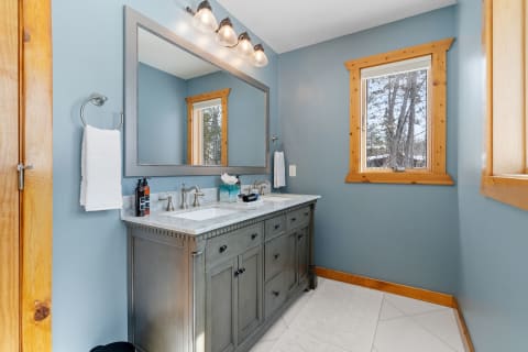 Modern bathroom with gray vanity, blue walls, natural light from window, and wooden accents.