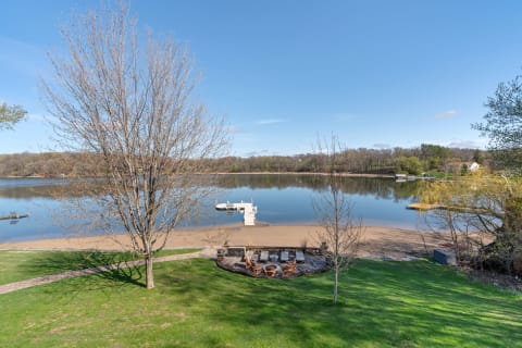 Lakeside view featuring a sandy beach, green lawn, and a dock under a blue sky.