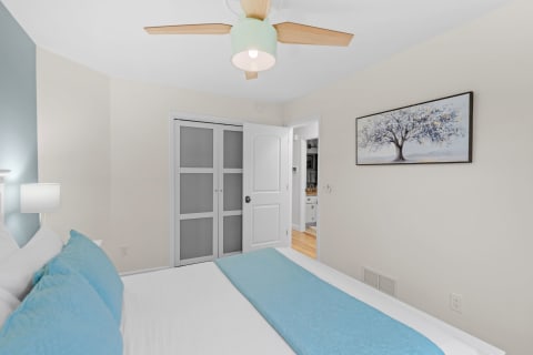 Bright and serene bedroom featuring a bed with white sheets and a turquoise runner, a ceiling fan, and wall art.