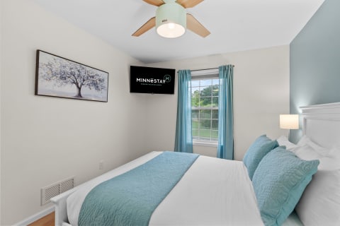 A cozy bedroom featuring a bed with turquoise accents, a tree painting, and a mounted TV.