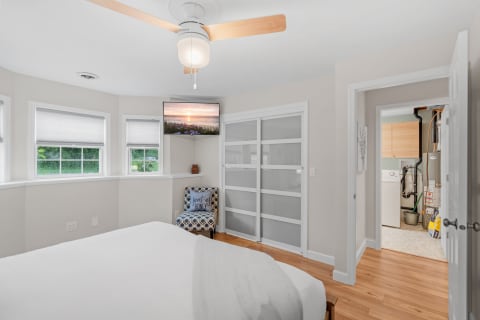 A bright bedroom with a bed, ceiling fan, TV showing a sunset, and a chair in the corner.