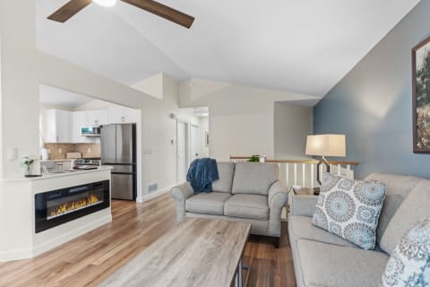 A modern living room with gray sofas, a coffee table, and an electric fireplace visible; an open kitchen is in the background.