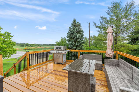 A wooden deck featuring a grill and outdoor seating, surrounded by greenery under a bright sky.