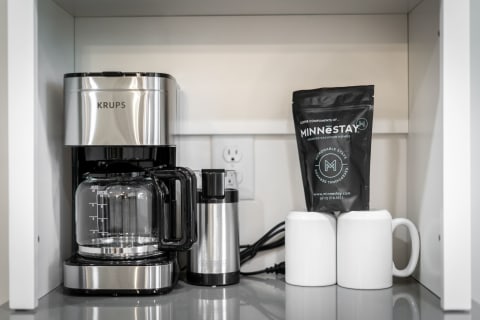 A modern coffee station with KRUPS coffee machine, frother, MINNESTAY coffee bag, and white mugs.