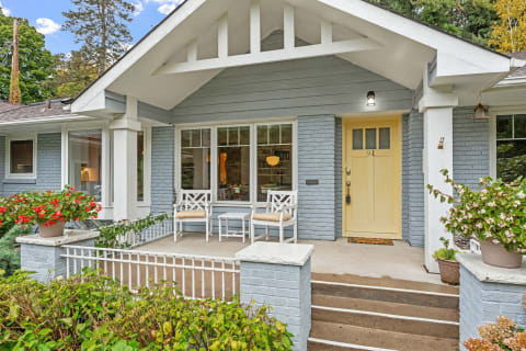 Front view of a charming house with a light blue facade, yellow door, and white porch chairs surrounded by flowers.