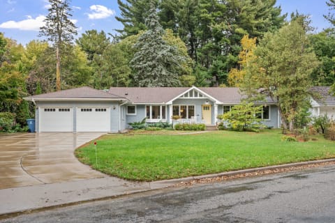 A light blue single-story house with a brown roof and two-car garage in a lush green setting.