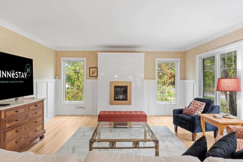 A living room featuring an ivory sofa, glass coffee table, and a flat-screen TV with a logo, alongside large windows with views of greenery.