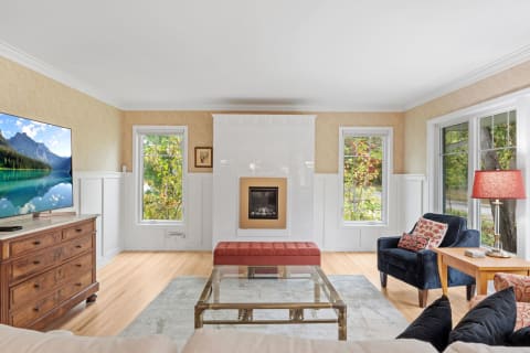 A contemporary living room with a white fireplace, plush ottoman, and large windows letting in sunlight.