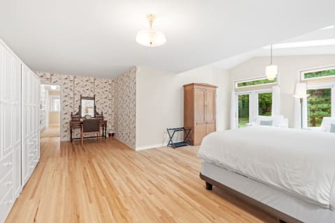 Spacious bedroom featuring light wood flooring, white closet doors, floral wallpaper, vintage vanity, and large windows.