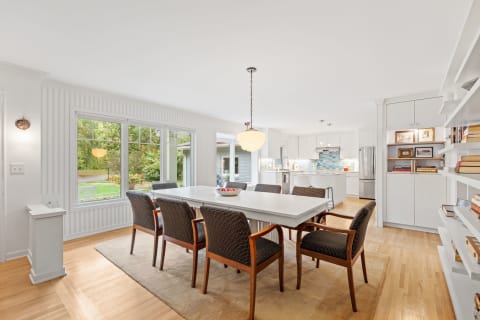 A modern dining area with a large table and stylish chairs, featuring windows with a view of greenery.