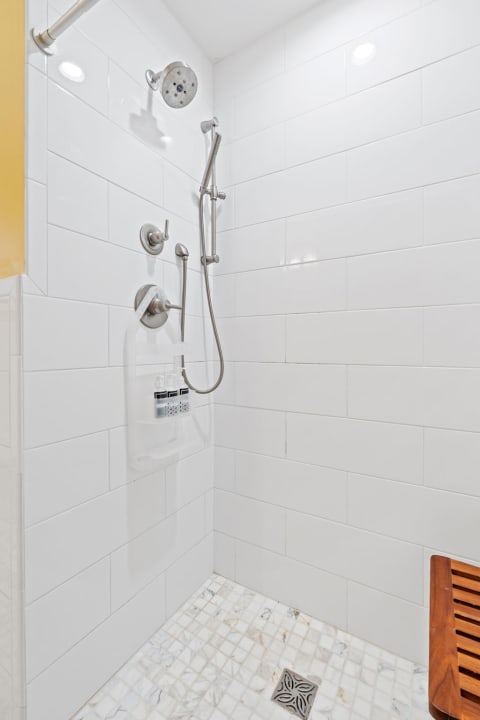 Shower area featuring white tiles, chrome fixtures, and a wooden bench.